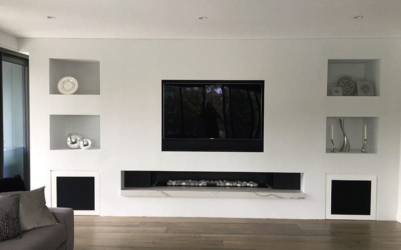 Home theatre wall with a recessed TV, in-wall speakers and a linear fireplace below