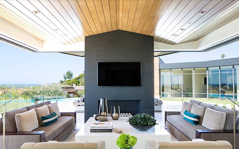 Television mounted on a dark feature chimney in an open-sided alfresco pavilion, with the pool and the water beyond