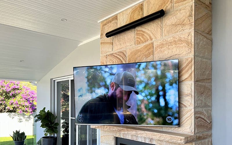 TV wall mounted on a sandstone chimney with an outdoor speaker bar above, in an alfresco area