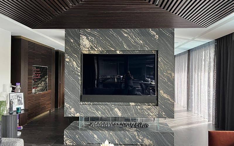 Large TV set into a black marble feature wall above a fireplace, under a slatted timber ceiling