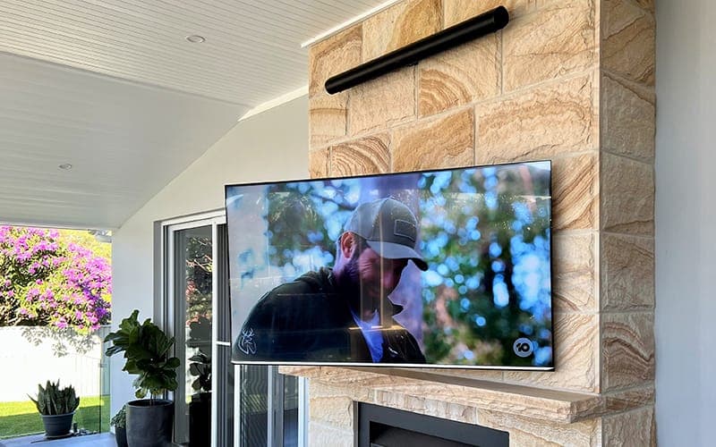 TV wall mounted on a sandstone chimney breast with a soundbar above, in a covered alfresco area